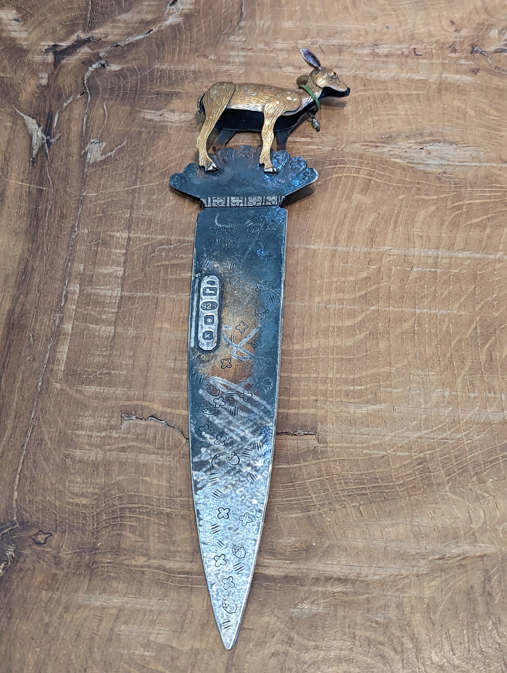 A fawn coloured enamelled silver deer soldered on the top of an oxidised silver paper knife. The knife has a silver tip and visible hallmarks. The deer has a green collar with a bell. 