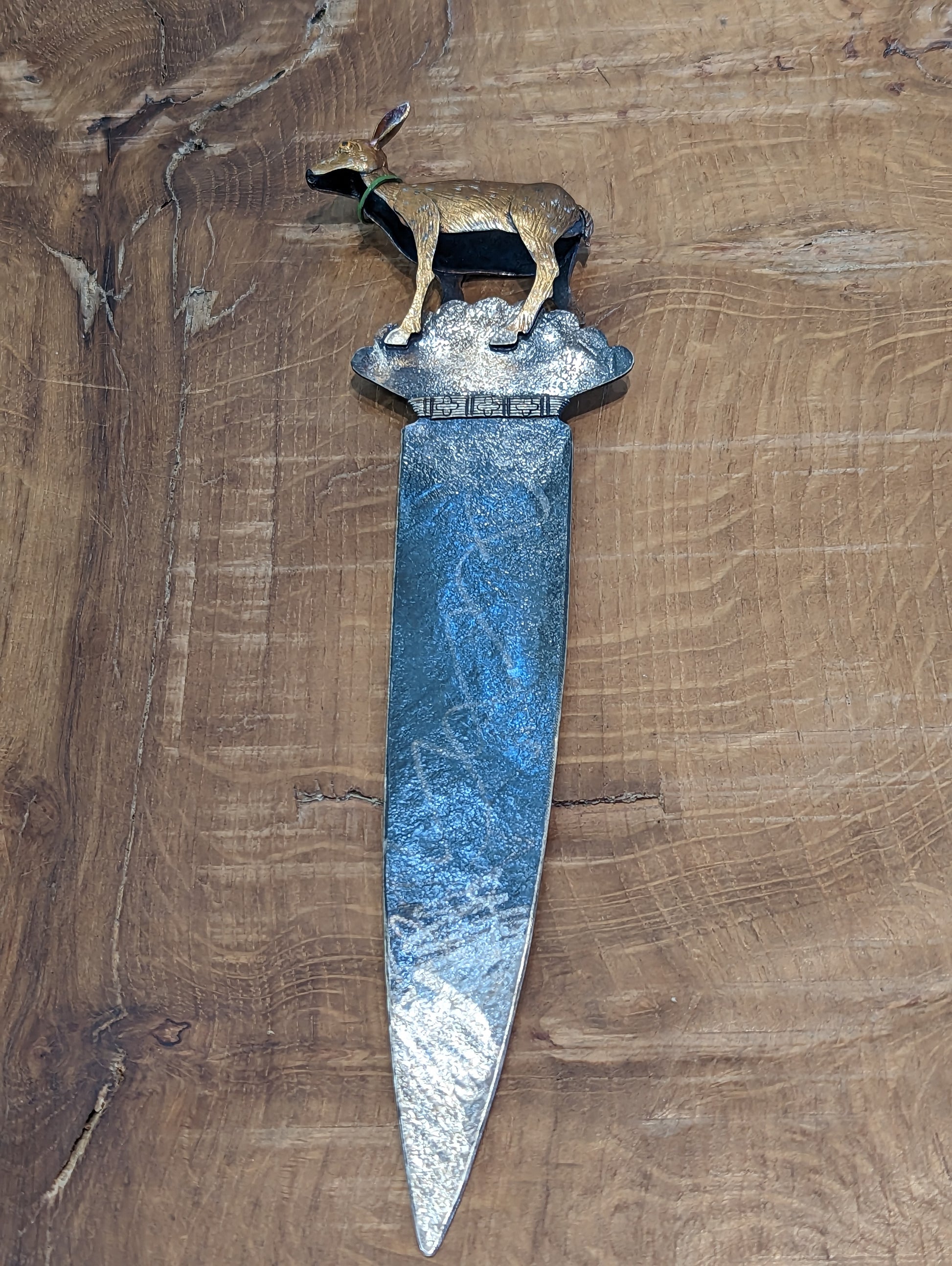 A fawn coloured enamelled silver deer soldered on the top of an oxidised silver paper knife. The knife has a silver tip. The deer has a green collar with a bell.