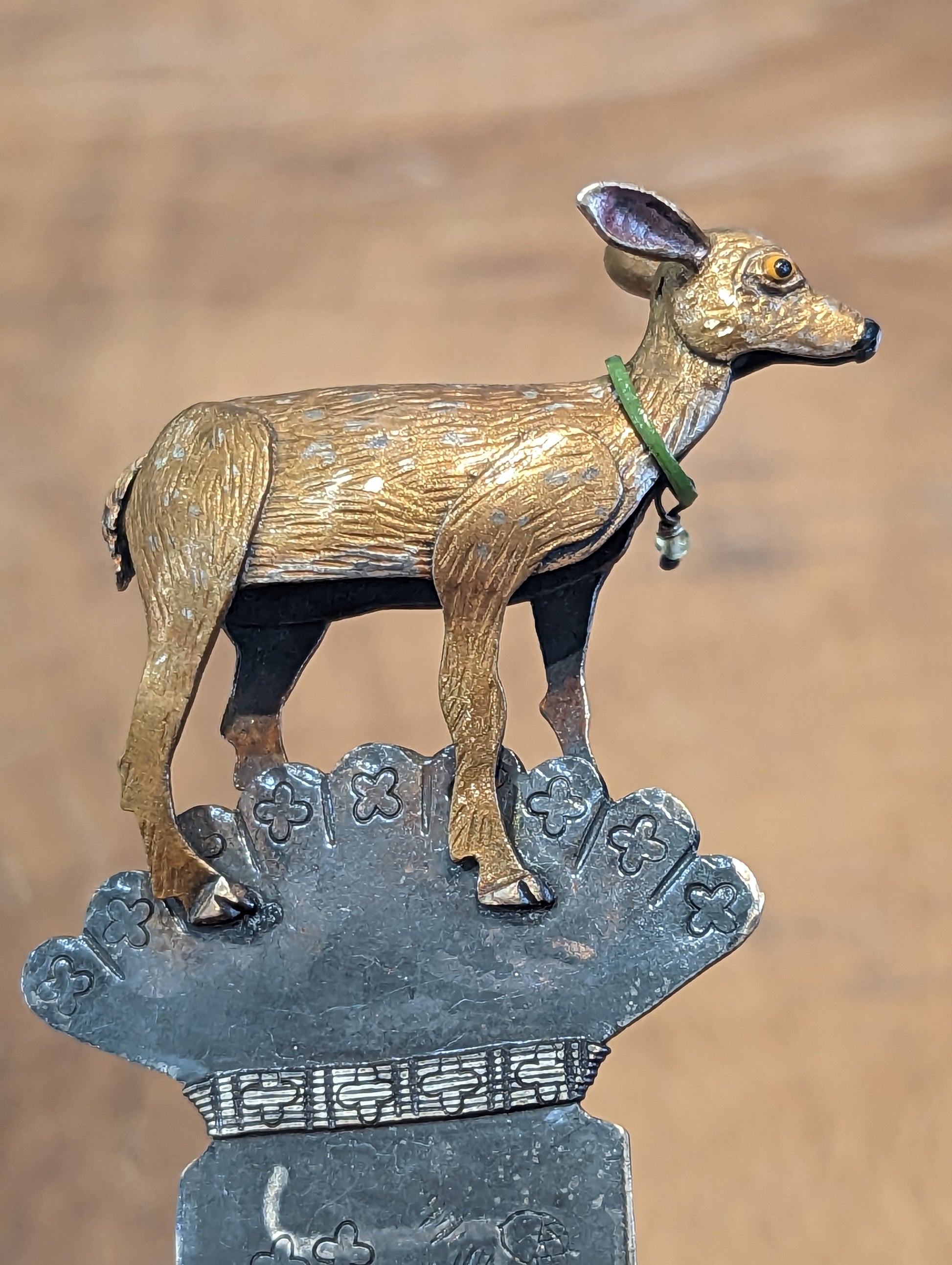 Close up of deer facing to the right. The deer has a green collar and yellow eyes. The deer in on top of a silver fan shape ornamentation. The leaves of the fan are decorated with four petalled flowers. 