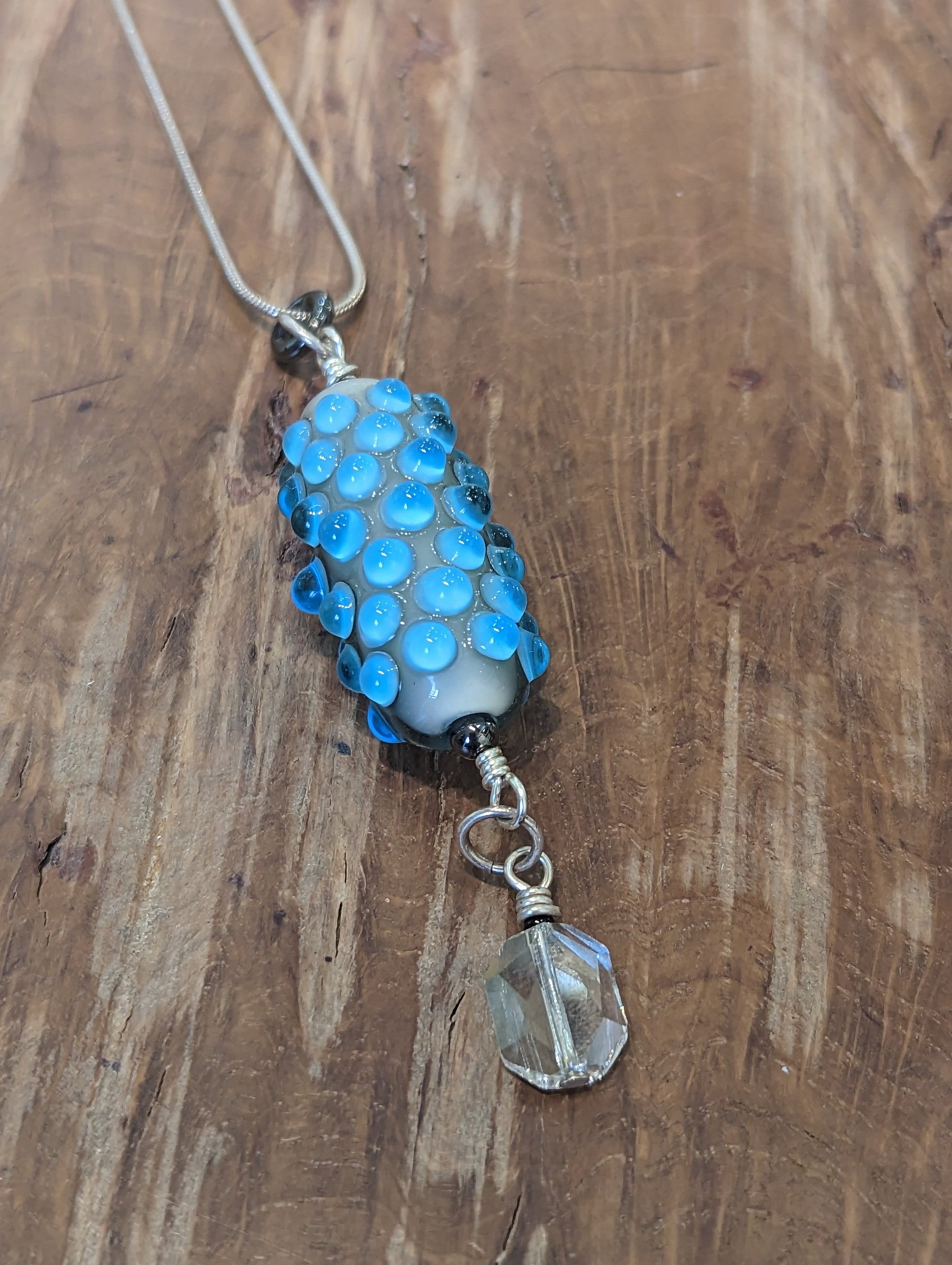 A blue glass pendant with lighter blue dots. It has a swarovski crystal dangling from it. 