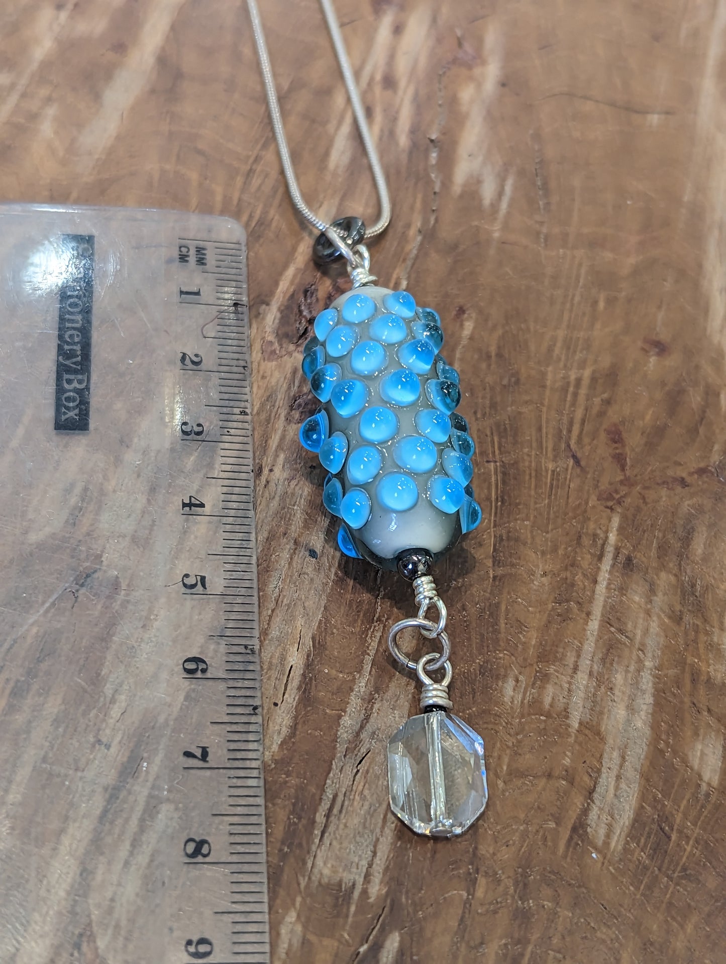 A blue glass pendant with lighter blue dots. It has a swarovski crystal dangling from it. The pendant is next to a ruler to demonstrate that it is 9cm long.  