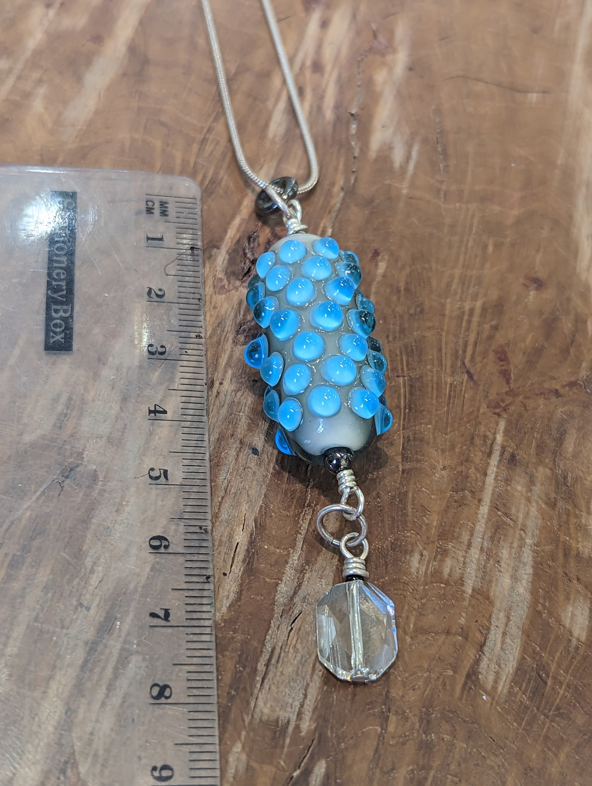 A blue glass pendant with lighter blue dots. It has a swarovski crystal dangling from it. The pendant is next to a ruler to demonstrate that it is 9cm long.  