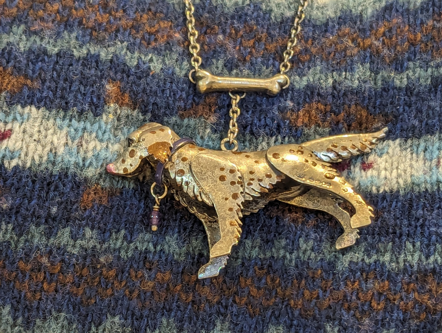 Side view of a silver dog pendant on top of a blue fair isle jumper. . The dog is facing to the left. It is silver with brown enamel spots. It has a blue collar with a purple bead dangling from it. 