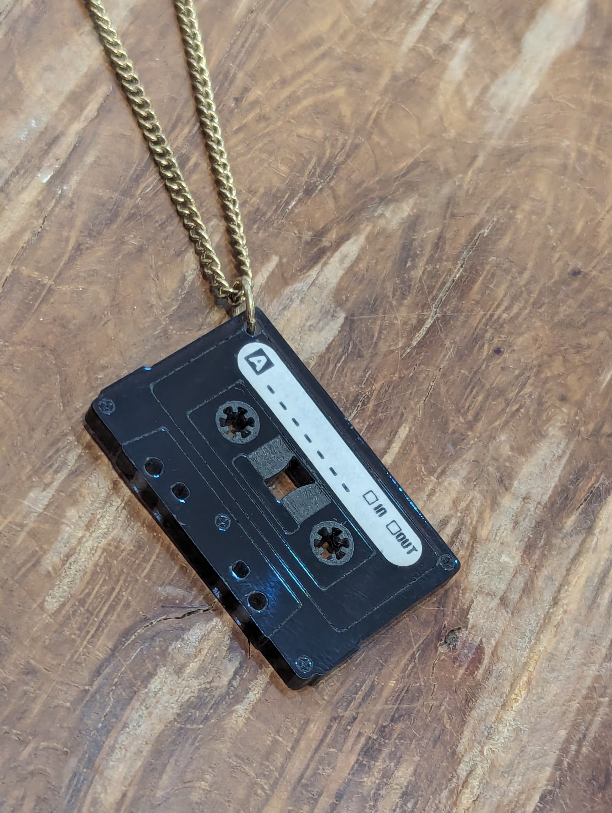 Black acrylic cassette tape pendant with etched details and a white label marking the a side. 