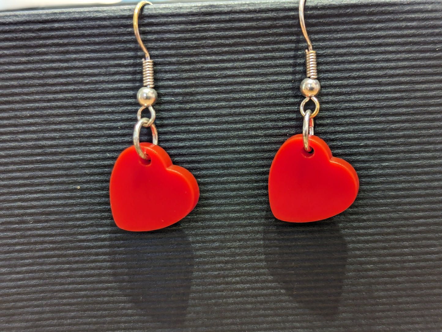 Two bright red hearts on silver earring hooks on a striped black background. 