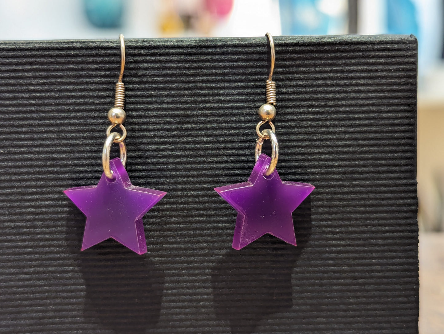 Pair of bright purple earrings in the shape of stars. Earrings are on silver hooks. They hang in front of a black background. 