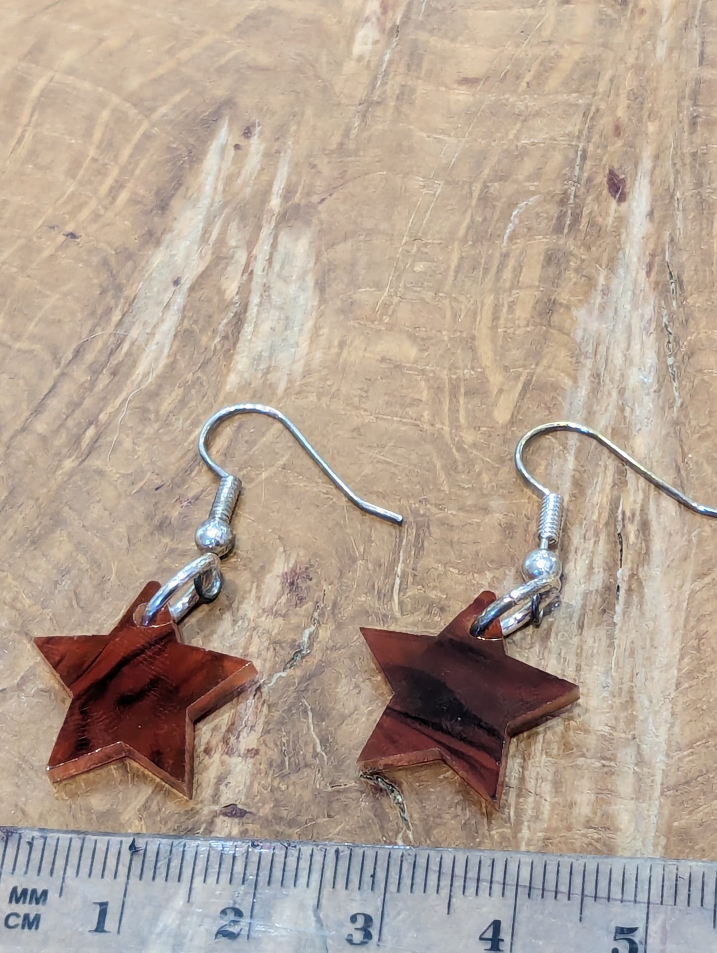 Brown Tortoiseshell star shaped earrings with silver hooks on a warm wooden background. They lie next to a ruler which shows that each star is 1.5cm in diameter.